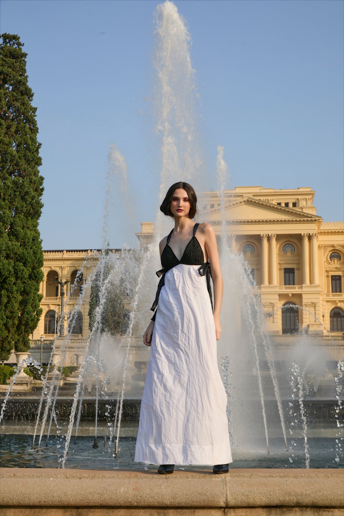 Vestido longo preto e branco da marca Isabela Monteiro. Decote realçado pelo bojo preto feito à mão em crochê. Costas nuas com decote de fitas cruzadas em laço nas laterais. Saia longa branca em algodão e inox para efeito amassado descontraído. Bolsos nas costuras laterais. 